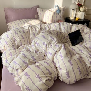 Peut inclure: Un lit avec un drap et des taies d'oreiller lavande. La housse de couette et les taies d'oreiller ont un fond blanc avec des rayures verticales lavande et des motifs de nœuds. Une tablette noire est posée sur le lit.