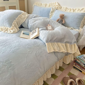 May include: A light blue bed set with a floral embossed pattern, featuring a duvet cover and pillows with cream-coloured ruffles. A small teddy bear and a perfume bottle rest on the bed. An open book and a stack of books are also visible.
