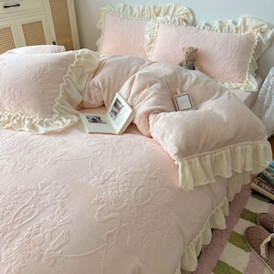 May include: A light pink bedding set with floral embroidery, featuring a comforter, pillows, and a bedspread. The set includes ruffled edges in a cream color. A small teddy bear and a book are on the bed.