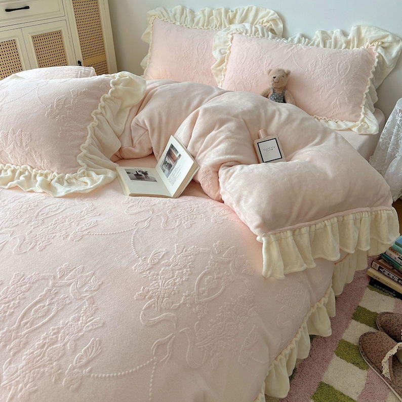 May include: A light pink bedding set with floral embroidery, featuring a comforter, pillows, and a bedspread. The set includes ruffled edges in a cream color. A small teddy bear and a book are on the bed.