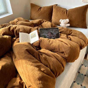 Może przedstawiać: Łóżko z brązową pościelą, w tym kołdrą i dwiema poduszkami. Otwarta książka i tablet leżą na łóżku. Mały biały miś siedzi na poduszkach. Pokój ma okno z żaluzjami.