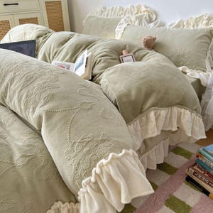 May include: A sage green bedspread with a floral pattern and ruffled edges. The bedding includes a comforter, pillows, and a bolster pillow. A small teddy bear and a perfume bottle are on the bed. The bed is made up with a soft, plush material.