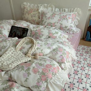 May include: A bed covered with a floral patterned duvet and pillows. The bedding features a white background with pink and green floral designs. A matching tote bag and a rug with a similar pattern are also visible.