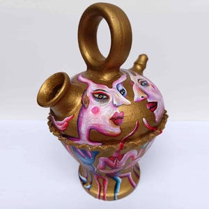 May include: A gold ceramic pitcher with a unique handle and painted with whimsical faces. The faces are pink and white with black eyes and red lips. The pitcher is decorated with gold and blue accents.