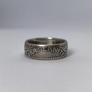 Puede incluir: Un anillo de plata con la palabra "FRATERNITE" grabada en la banda. El anillo tiene una superficie texturizada y un aspecto vintage. El anillo es una joya.