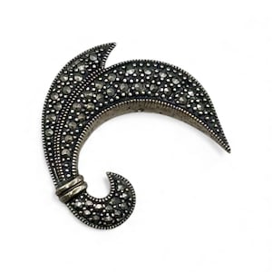 May include: A vintage silver-tone brooch in a stylized leaf design. The brooch is encrusted with small, round, dark gray stones, creating a textured surface. The design features a curved shape with a pointed tip and a looped base.