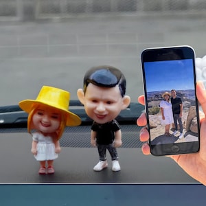 Puede incluir: Dos figuras de bobblehead en miniatura de una pareja, una con sombrero amarillo y vestido blanco, la otra con camisa negra y pantalón gris. Un smartphone muestra una foto de la pareja en un entorno exterior pintoresco.