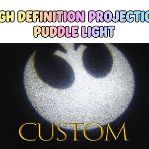 Op de afbeelding: Een aangepaste plaslichtprojectie van een wit symbool op een donkere ondergrond. De tekst "HIGH DEFINITION PROJECTION PUDDLE LIGHT" staat bovenaan en "CUSTOM" onderaan.