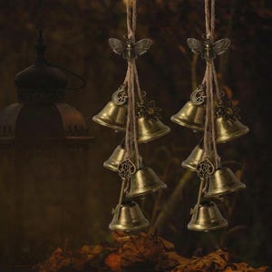 May include: Two bronze-colored wind chimes with small bells hanging from twine. Each chime features a butterfly at the top, with sun, moon, and key charms. The background is a blurred, dark brown with a lantern.