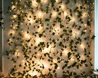 May include: A decorative string of warm white LED lights intertwined with artificial ivy vines. The green leaves and small lights create a soft, ambient glow, perfect for home decor or event styling. The lights are evenly spaced along the vines.