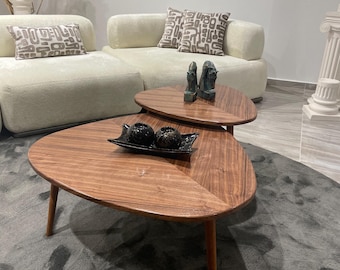 Puede incluir: Juego de dos mesas de centro de madera con forma orgánica. Las mesas tienen un rico acabado de vetas de madera y están soportadas por patas delgadas y cónicas. Objetos negros decorativos se encuentran en la mesa superior. Un sofá y cojines a juego están en el fondo.