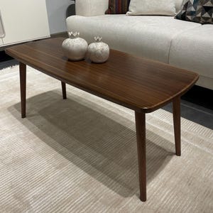 Puede incluir: Una mesa de centro rectangular de madera marrón oscuro, de estilo moderno de mediados de siglo. Tiene cuatro patas cónicas y esquinas redondeadas. Dos jarrones decorativos de cerámica blanca con detalles de corona están sobre la mesa. La mesa está sobre una alfombra clara.