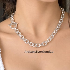 Puede incluir: Un collar de cadena de eslabones plateados con un cierre de barra prominente. El collar consta de eslabones metálicos redondeados y uniformes conectados en un bucle continuo, con el texto ArtisansilverGoodCo visible debajo de la cadena.