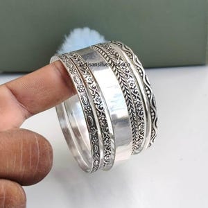 May include: A set of silver bangles featuring a mix of smooth polished bands and bands with intricate floral and braided textures. The text ArtisansilverGoodCo is visible on the central smooth band of the silver bracelet stack.