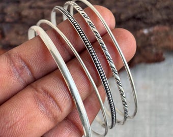 Peut inclure: Un assortiment de bracelets argentés, chacun avec un design unique. Les bracelets présentent des textures variées, certains avec des surfaces lisses, tandis que d'autres affichent des motifs complexes de points et de torsades. Les bijoux sont présentés sur un fond en bois naturel.