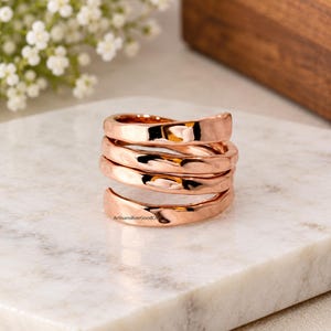 Peut inclure: Une bague couleur or rose présentant un design en spirale à plusieurs niveaux avec une surface polie et ondulée. L'anneau s'enroule sur lui-même plusieurs fois, créant un aspect empilé avec des courbes lisses et réfléchissantes.