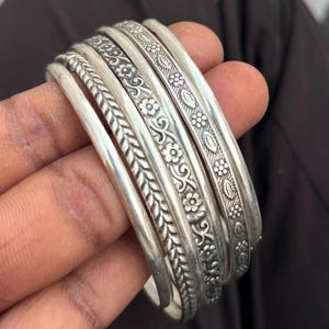 Könnte beinhalten: Eine Sammlung silberner Armreifen mit aufwendigen Blumen- und Blattmustern. Die Armreifen sind gestapelt und zeigen die detaillierten Designs. Das Metall hat eine polierte Oberfläche, die das Licht reflektiert und die geprägten Texturen hervorhebt.