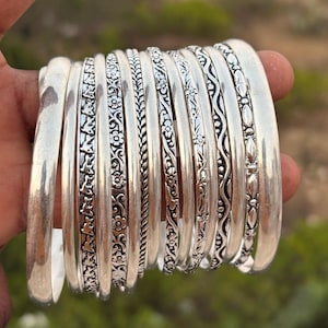 Op de afbeelding: Een verzameling zilveren armbanden met diverse decoratieve patronen, waaronder bloemen- en geometrische ontwerpen. De armbanden worden bij elkaar gehouden, wat hun gepolijste afwerking en ingewikkelde details benadrukt. Geschikt voor dagelijks gebruik of speciale gelegenheden.