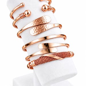 Puede incluir: Una colección de cinco brazaletes de metal en tono oro rosa exhibidos en un soporte cilíndrico blanco. El conjunto incluye un brazalete con extremos de bola, uno con textura martillada, dos brazaletes lisos pulidos y uno con diseño de malla.