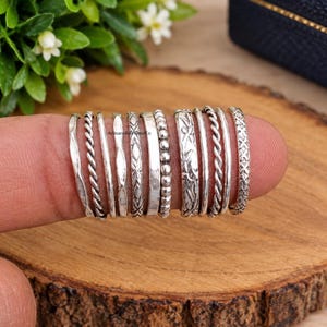 Puede incluir: Una colección de once anillos de plata mostrados en un dedo, con diversas texturas que incluyen patrones de cuerda trenzada, cuentas, lisos y grabados geométricos. Los anillos están dispuestos lado a lado, mostrando una variedad de acabados.