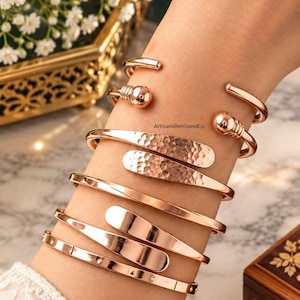 May include: A set of five rose gold-toned cuff bracelets worn on a wrist. The bracelets feature various designs, including a ball-end cuff, a hammered texture cuff, a smooth polished cuff, a tapered cuff, and a thin cuff with small dot accents.