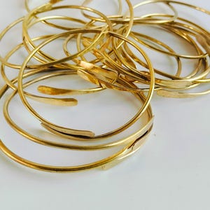 May include: A collection of gold-colored bangles, some with a hammered texture, are arranged on a white surface. The bangles vary in design, with some featuring a simple, rounded shape and others having a more flattened, rectangular element.