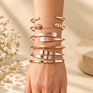 May include: A stack of five rose gold-toned metal bracelets worn on the wrist. The set includes a cuff with ball ends, a hammered texture cuff, a smooth polished cuff, a flat-ended cuff, and a thin band with small circular indentations.