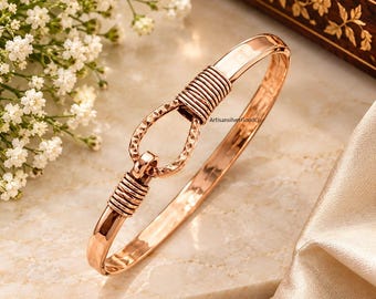 May include: A rose gold-toned bangle bracelet featuring a central oval-shaped link with a textured, faceted surface. The bracelet is accented by two sections of coiled wire detailing on either side of the central link, creating a polished, metallic finish.