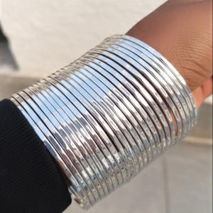 May include: A stack of silver bangles on a wrist. The bangles are thin, shiny, and have a hammered texture. The wrist is wearing a black sleeve. The background is out of focus.