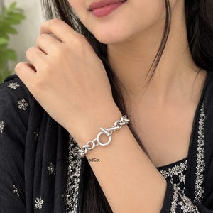 May include: A silver-toned chain link bracelet featuring a prominent toggle clasp closure. The bracelet is composed of thick, interlocking circular links that create a continuous, uniform pattern around the wrist, secured by a T-bar and ring.