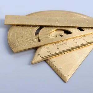 Puede incluir: Un conjunto de herramientas de dibujo de latón dorado que incluye una regla recta, un transportador semicircular y una escuadra triangular. Las herramientas tienen marcas de medición grabadas y el texto XWWJ-FDC STATIONERY en la escuadra.