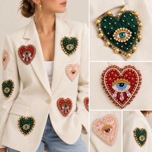 Puede incluir: Un blazer de color crema adornado con múltiples parches en forma de corazón. Cada parche presenta intrincados abalorios, detalles metálicos y un motivo de ojo central en rojo, verde o rosa. El blazer tiene solapa y un botón.