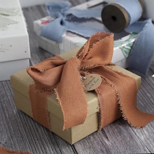 以下が含まれることがあります： テラコッタ色の幅広で端がほつれた布製リボンが蝶結びにされた、正方形の段ボール製ギフトボックス。リボンには「Handmade with love」という黒い文字が印刷された、小さな円形のベージュ色の紙タグが取り付けられています。