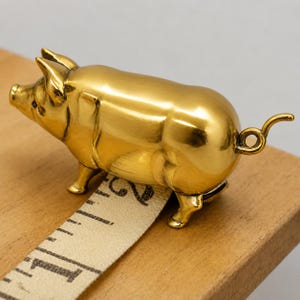 Puede incluir: Una figura de cerdo de metal dorado pulido con una cola enroscada y orejas puntiagudas. El cerdo está colocado sobre una cinta métrica blanca que muestra una medida de aproximadamente 5 cm, indicando la escala del pequeño objeto decorativo.