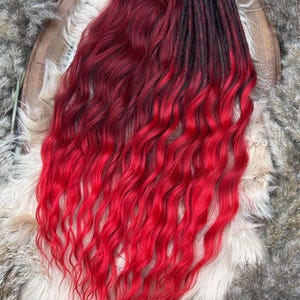Pode incluir: Um feixe de rastas vermelhas e pretas com uma textura ondulada. O cabelo transita de raízes escuras para um vermelho vibrante nas pontas. As rastas são exibidas em uma bandeja de madeira decorativa com um fundo fofo de tons neutros.