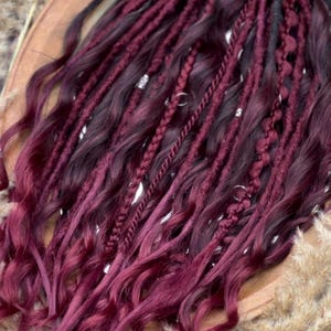 Puede incluir: Dreadlocks burdeos y negros con puntas onduladas. Algunos están trenzados y adornados con anillos plateados. El cabello está dispuesto sobre una superficie de madera con hierba seca en el fondo.
