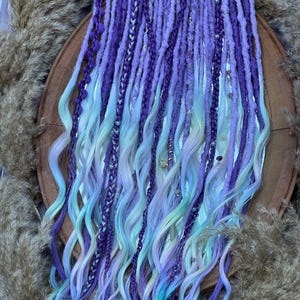 May include: A collection of purple, lavender, and blue dreadlocks. The dreads are arranged in a vertical pattern, with some sections braided. The ends of the dreadlocks transition to a lighter blue and green color. Some dreads are adorned with small beads.