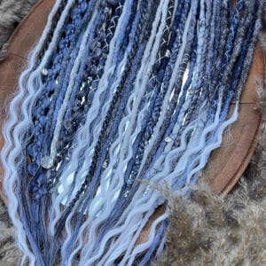 Puede incluir: Colección de rastas sintéticas azules, grises y blancas. Las rastas están dispuestas sobre una superficie de madera y presentan varias texturas, incluyendo trenzas y puntas onduladas. Algunas rastas están adornadas con detalles plateados.