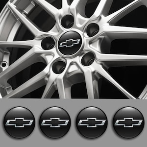 Op de afbeelding: Close-up van een zilveren lichtmetalen velg met een zwarte naafkap met een zilveren Chevrolet-logo. Hieronder worden vier extra zwarte naafkappen met hetzelfde logo weergegeven.