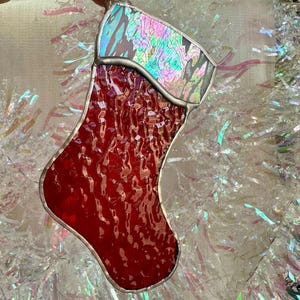 May include: A stained glass Christmas stocking ornament. The stocking is primarily red with a textured pattern and a silver border. The top of the stocking has an iridescent, multi-colored design. A silver loop is at the top for hanging.