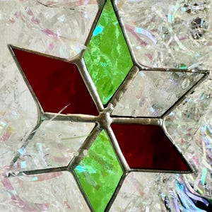 May include: A handcrafted stained glass star ornament. The star features red, green, and clear glass panes, with a silver-colored metal frame. The ornament is designed with a six-pointed star shape and is suspended by a silver loop.
