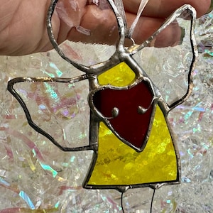 May include: Stained glass angel ornament with a yellow dress, red heart, and clear wings. The angel has a silver-colored metal frame and a ribbon for hanging. A decorative item for the holidays or year-round.