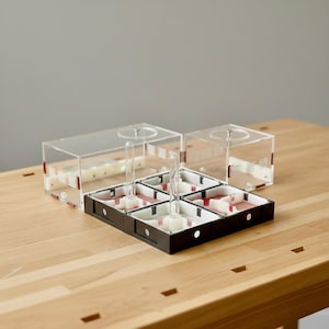May include: A clear acrylic ant farm with a black base and red accents. The farm has multiple compartments and a clear top, with two additional clear acrylic containers. The setup is on a wooden surface.