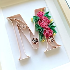 Può includere: Un'opera d'arte in quilling incorniciata con la lettera "M". La lettera è di un rosa tenue, con volute decorative. La lettera è decorata con rose rosa, foglie verdi e accenti gialli. L'opera d'arte è ambientata su uno sfondo bianco.