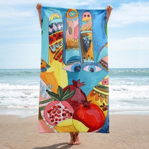 May include: A colorful beach towel with an abstract design featuring pomegranates, eyes, and geometric shapes in shades of blue, orange, red, and yellow. The towel is held up on a sandy beach with the ocean in the background.