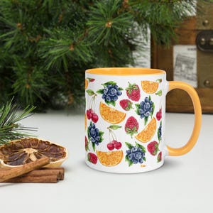 May include: White ceramic mug with a yellow interior and handle, decorated with a colourful pattern of blueberries, raspberries, cherries, and orange slices. The mug is set against a background of greenery and a wooden chest.