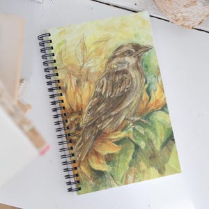 May include: A spiral-bound notebook featuring a watercolor illustration of a brown bird perched among yellow sunflower petals and green leaves. The cover art displays a detailed depiction of the bird with textured feathers and a soft yellow background.