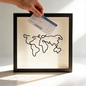 Op de afbeelding: Zwart omkaderde wandkunst met een minimalistische zwarte draadomtrek van een wereldkaart. Een hand houdt een instapkaart boven het kunstwerk. Het kunstwerk heeft een crèmekleurige achtergrond.