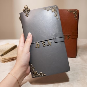 May include: A gray leather-like journal featuring antique brass-colored metal corner protectors, a fleur-de-lis accent, and a horizontal elastic band holding three dangling metal letter charms that spell out the name BEN in capital letters.