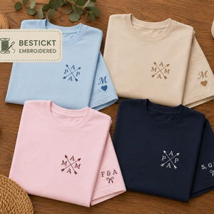 Könnte beinhalten: Vier gefaltete T-Shirts mit Rundhalsausschnitt in Hellblau, Beige, Hellrosa und Marineblau. Jedes Shirt zeigt auf der Brust gestickte gekreuzte Pfeile mit Buchstaben sowie personalisierten Text mit Symbolen wie Herzen oder Schleifen am Ärmelbündchen.